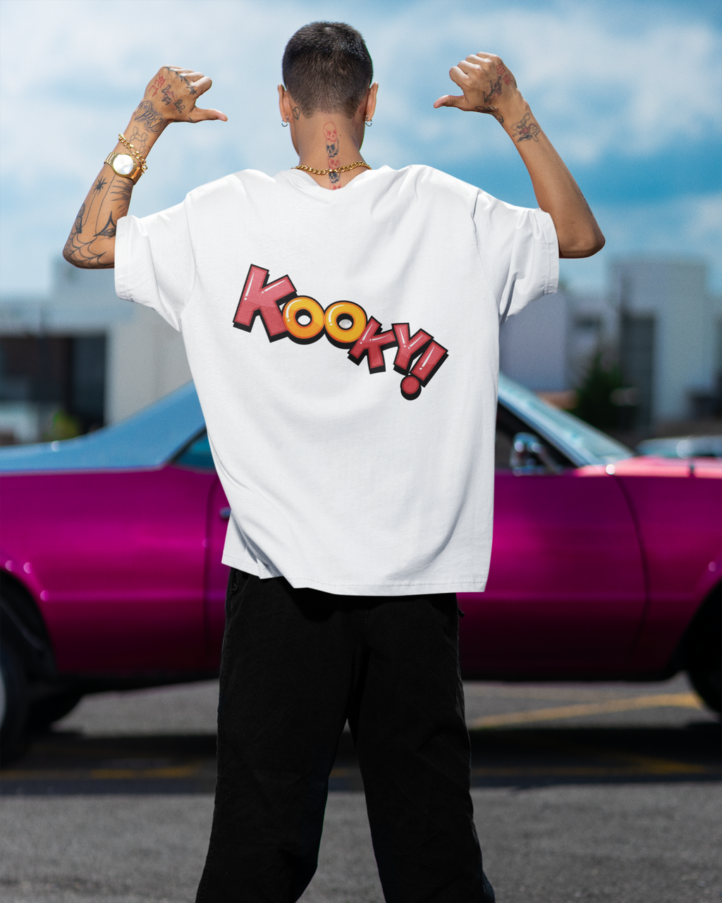 Person wearing a white t-shirt with 'Kooky!' design, standing in front of a pink car.