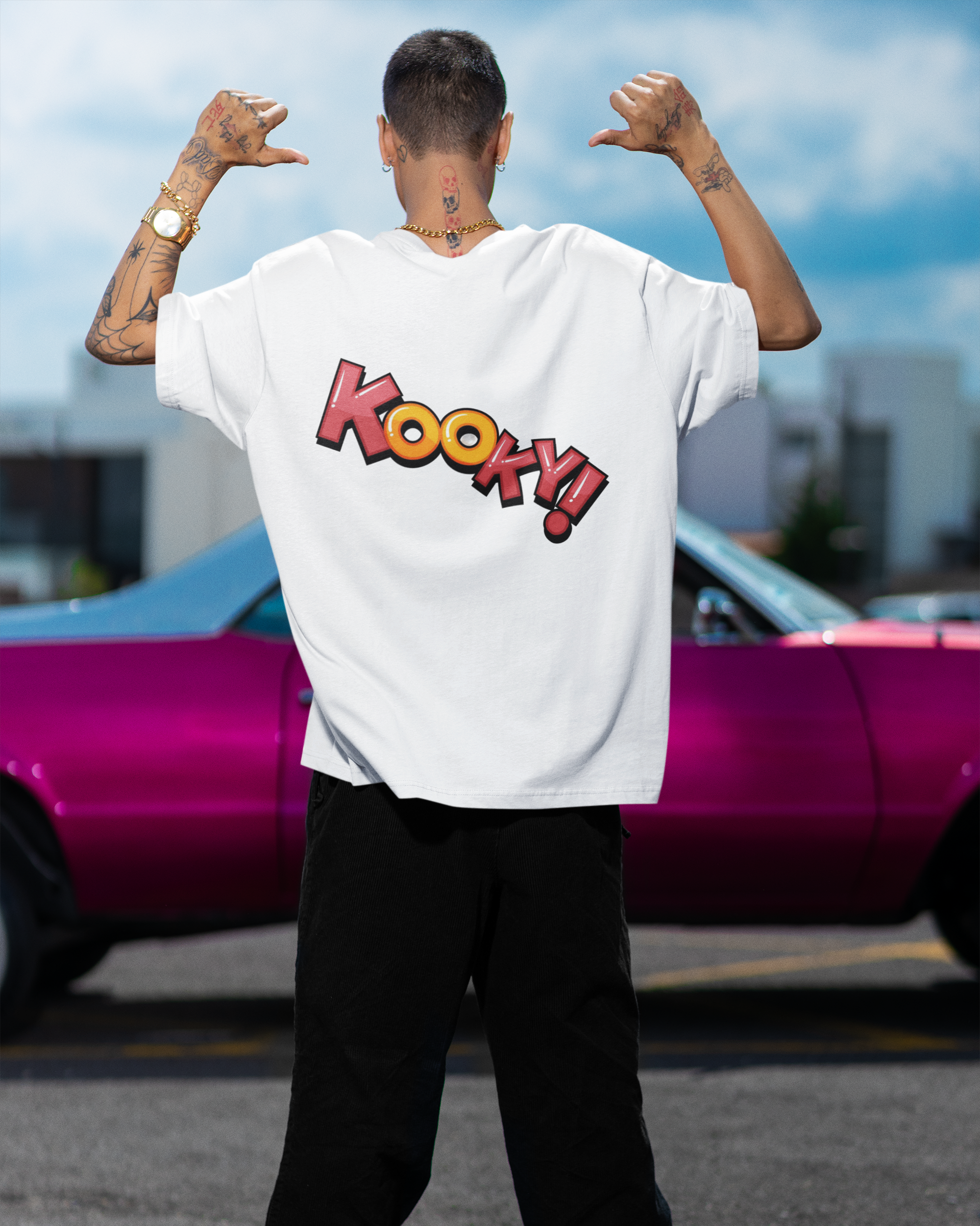 Person wearing a white t-shirt with 'Kooky!' design, standing in front of a pink car.