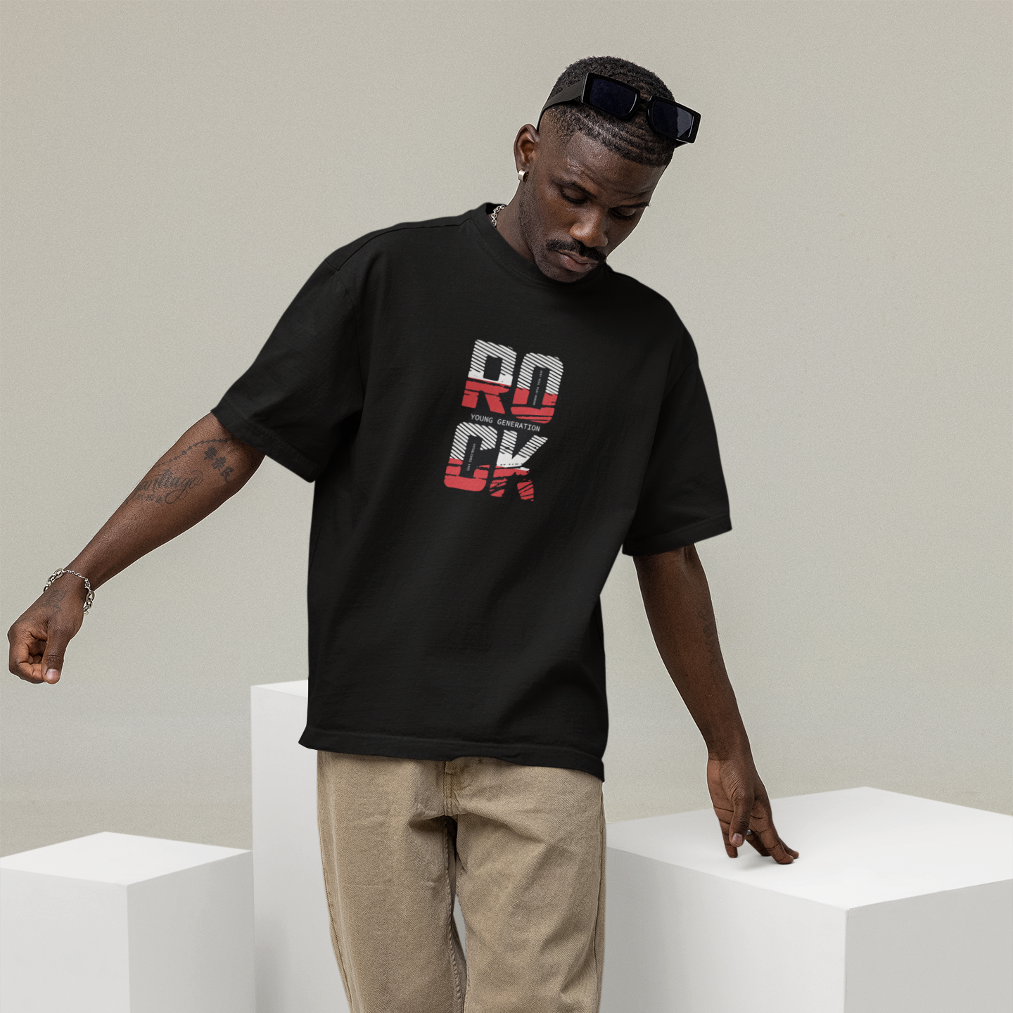 Man wearing a black t-shirt with 'ROCK' print, beige pants, and white sneakers in a minimalistic setting.