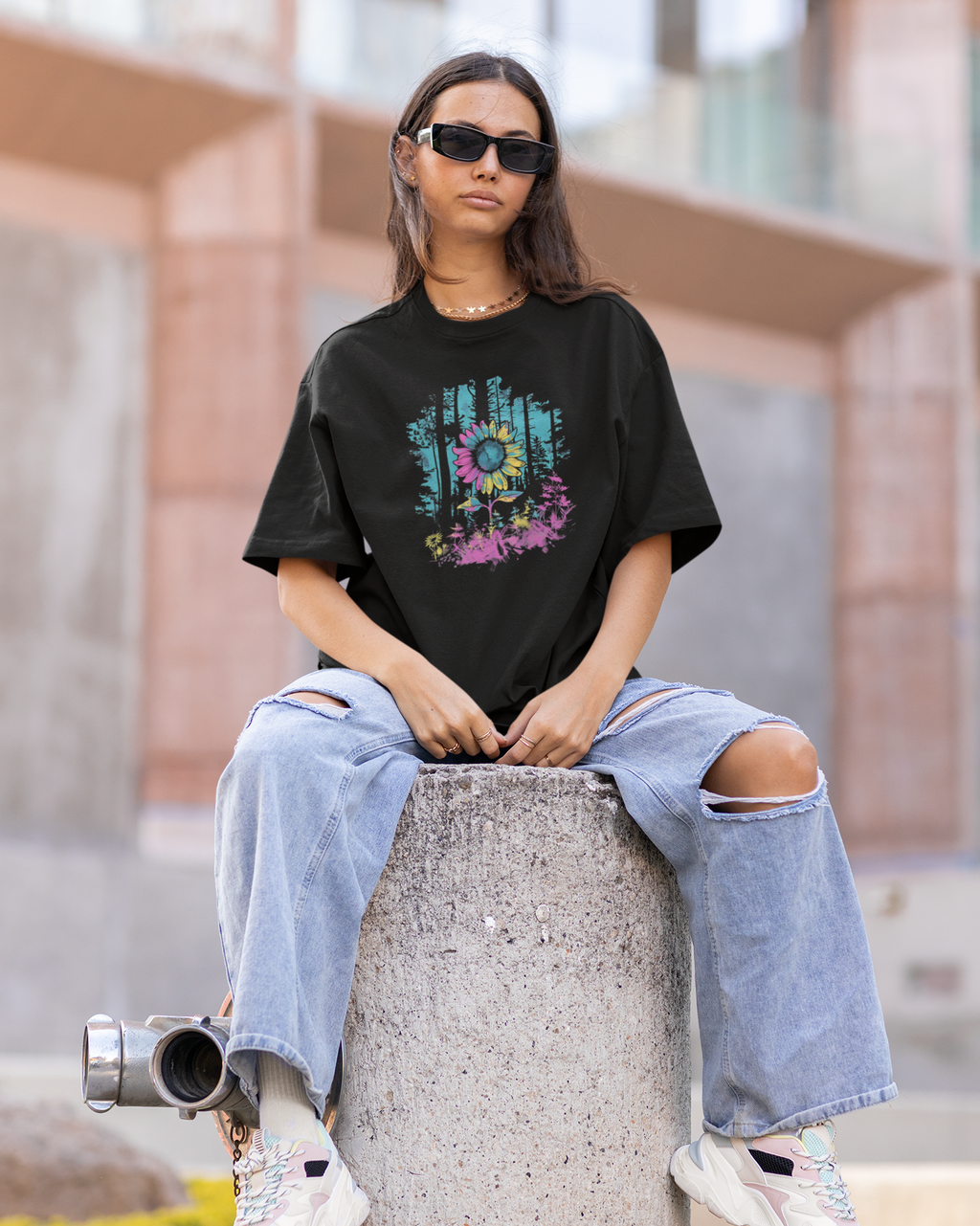 Person wearing a black t-shirt with colorful design and ripped jeans sitting on a concrete block.